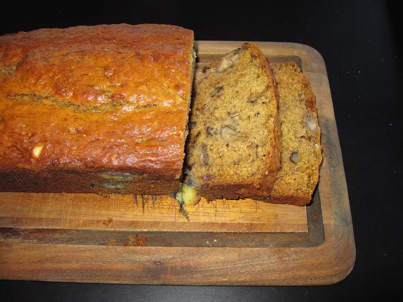 Golden banana bread loaf on a wooden board with two thick slices cut, showing a moist, walnut-studded crumb