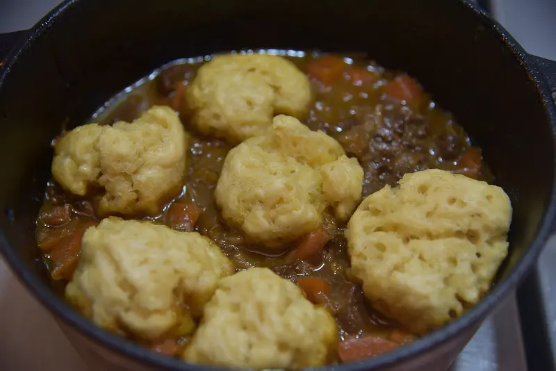 Fluffy suet dumplings sitting on a rich meat and carrot stew in a cast-iron casserole pot