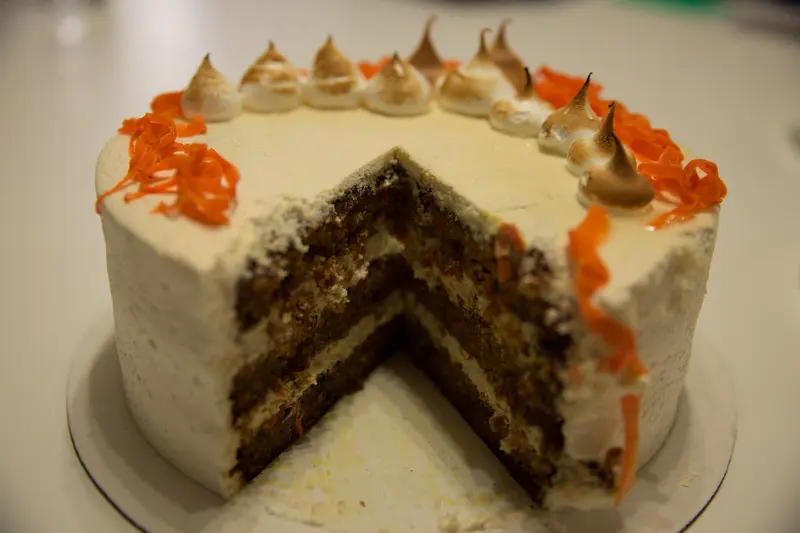 Layered carrot cake with cream cheese frosting and a slice cut away, decorated with shredded carrot and meringue kisses