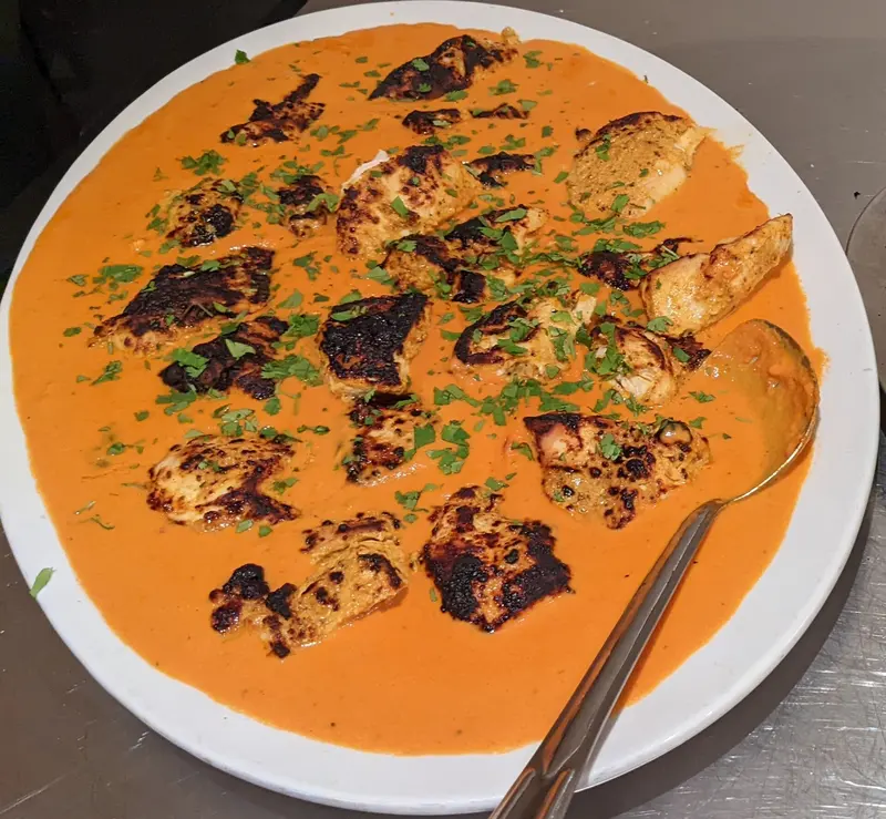 Charred chicken tikka pieces in a creamy orange tomato masala sauce, scattered with coriander, on a white plate