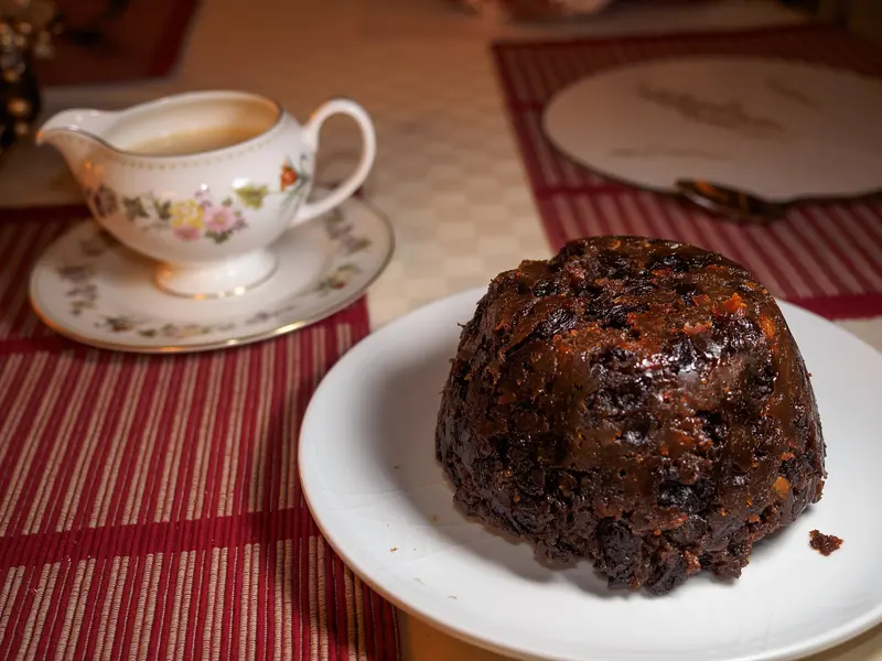 Whole dark, glossy Christmas pudding on a white plate with a jug of custard behind