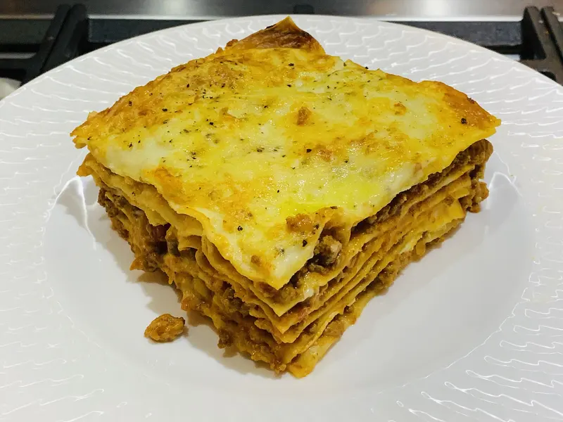 Square slice of beef lasagne showing layers of pasta and meat ragu under a golden baked top, on a white plate
