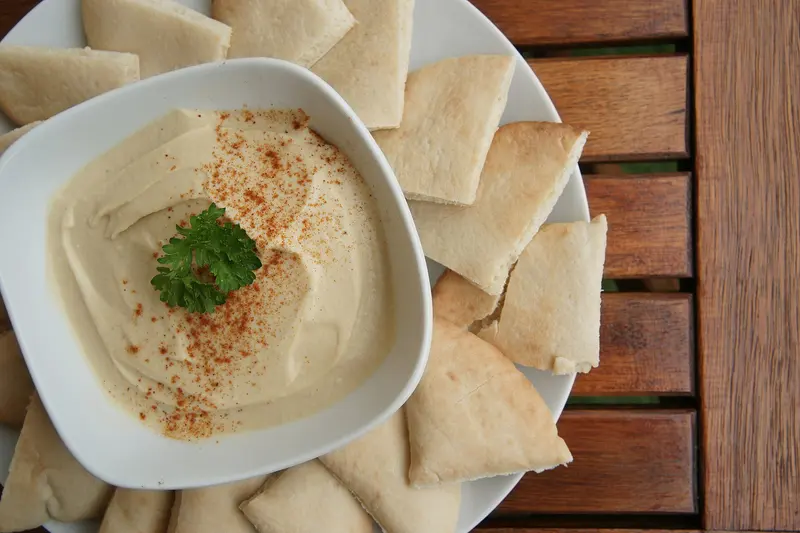 Bowl of smooth hummus dusted with paprika and topped with parsley, surrounded by pitta bread triangles