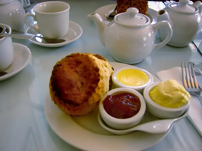 Golden scone on a plate with small pots of strawberry jam, clotted cream and butter, with teapots behind