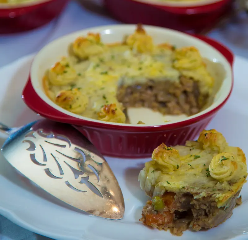 Shepherd's pie in a red baking dish with a serving scooped out, showing lamb mince, carrots and peas under piped golden mash