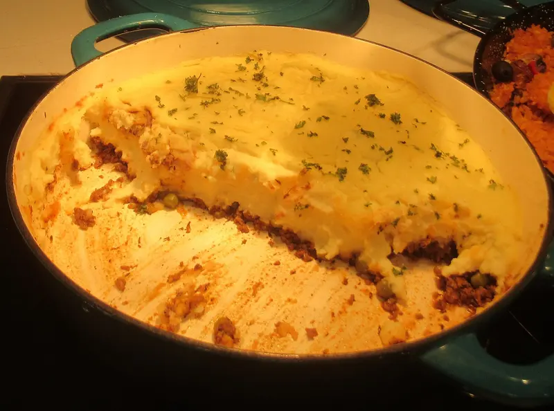 Cottage pie in a green cast-iron casserole, golden mashed potato top sprinkled with parsley over beef mince and peas