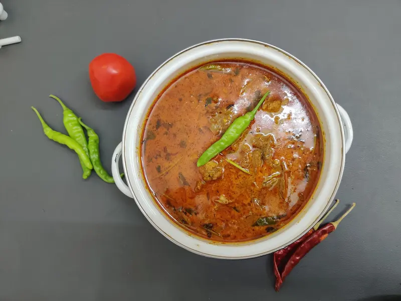 Overhead view of chicken curry in a rich spiced gravy in a white pot, garnished with green chilli, beside a tomato and chillies