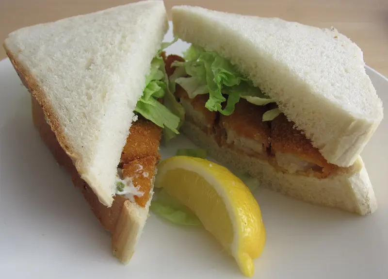 Fish finger sandwich on soft white bread with lettuce and tartare sauce, cut into triangles with a lemon wedge