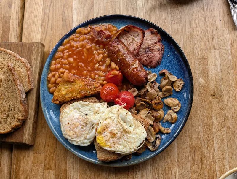 Overhead view of a full English breakfast on a blue plate: sausage, bacon, fried eggs, beans, mushrooms, tomatoes and toast