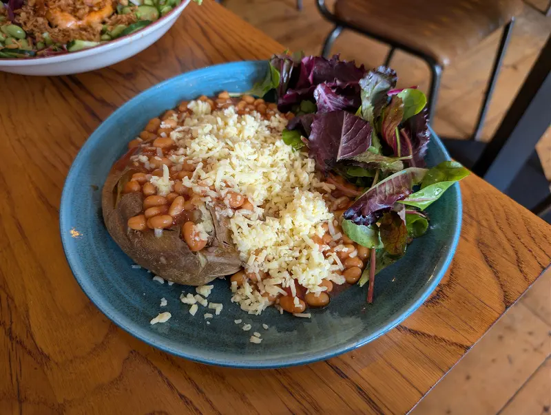 Jacket potato topped with baked beans and grated cheddar, with a side of mixed salad leaves on a blue plate