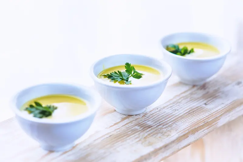 Three small white bowls of smooth, creamy leek and potato soup garnished with olive oil and parsley on a wooden board