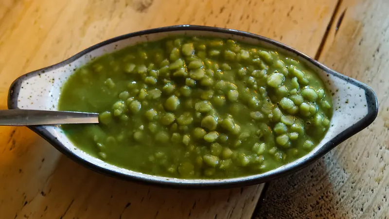 Bright green mushy peas in an oval speckled serving dish with a spoon, on a wooden table