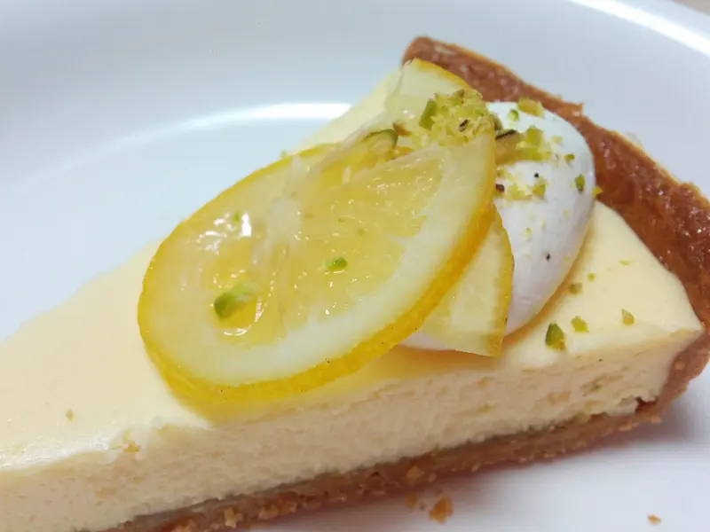 Slice of creamy lemon cheesecake on a biscuit base, topped with lemon slices, cream and chopped pistachio