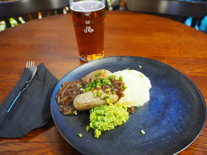 Glossy caramelised onion gravy spooned over a sausage with mashed potato and mushy peas on a dark blue plate