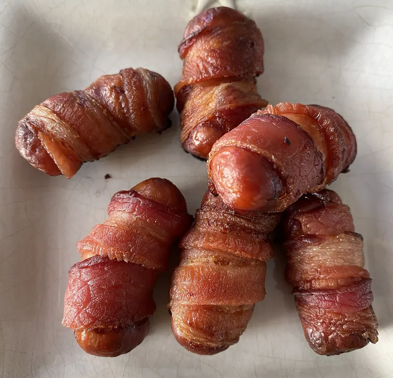 Roasted pigs in blankets: chipolata sausages wrapped in crisp, browned streaky bacon on a white plate