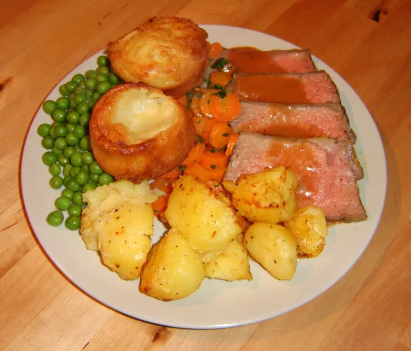 Sunday roast plate of pink sliced roast beef with gravy, two Yorkshire puddings, roast potatoes, carrots and peas