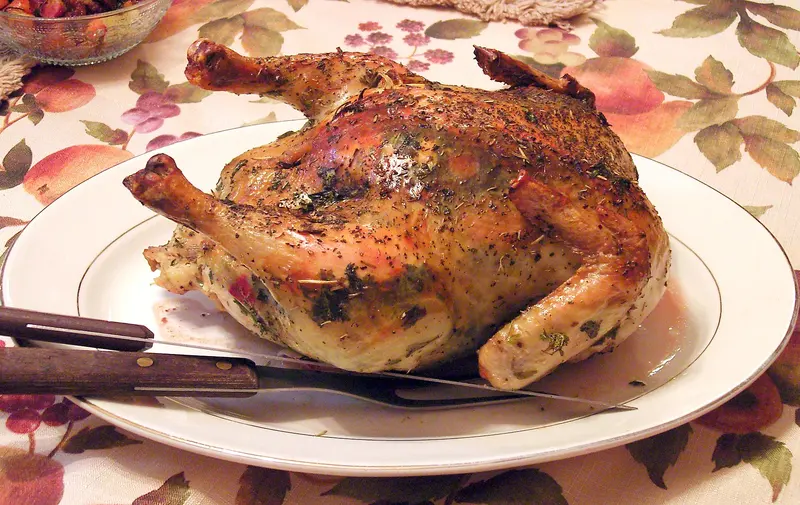 Golden whole roast chicken sprinkled with herbs on a white platter, with a carving knife and fork in front