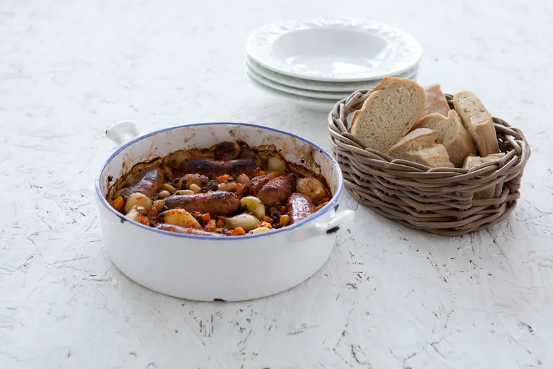Sausage and bean casserole in a white enamel dish, served with a basket of sliced crusty bread