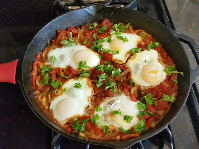 Shakshuka in a cast-iron skillet with five fried-set eggs in a chunky tomato and pepper sauce, scattered with parsley