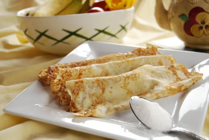 Thin golden British pancakes rolled up on a white plate, with a spoonful of sugar beside them
