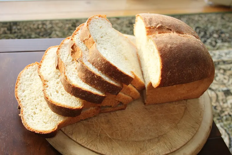 Golden-crusted white tin loaf partly sliced on a round wooden breadboard, showing its soft white crumb