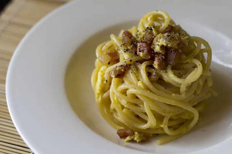 Twirled nest of spaghetti carbonara in a glossy egg and cheese sauce, topped with crisp cubes of pork, in a white bowl