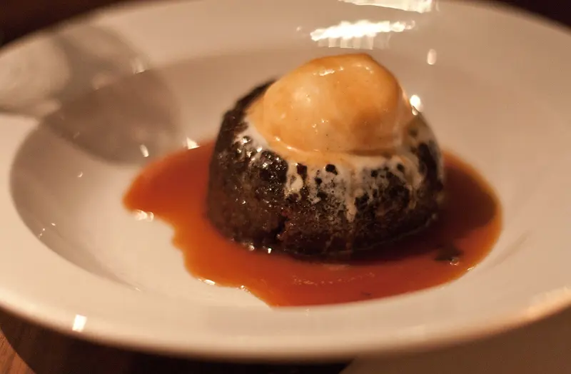 Dark sticky toffee pudding sitting in a pool of glossy toffee sauce, topped with a scoop of ice cream in a white bowl