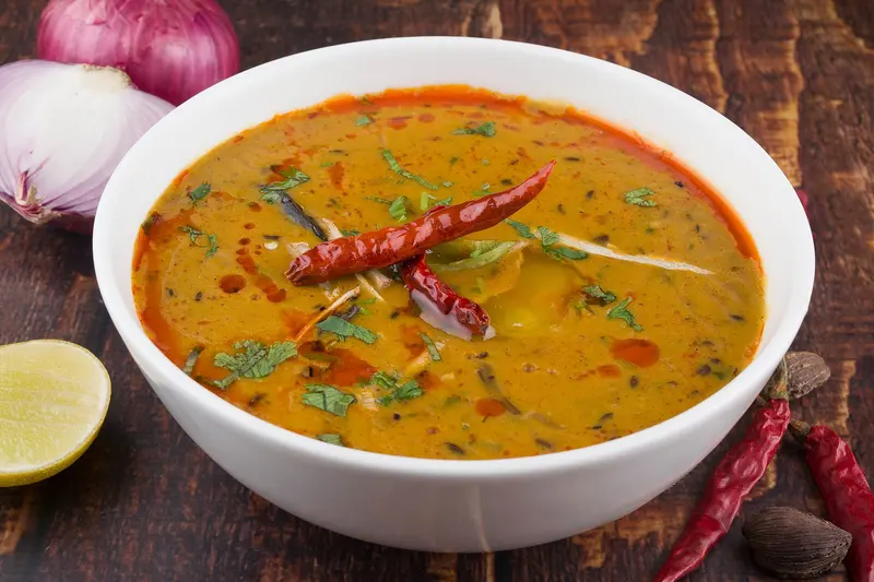 White bowl of golden tarka dal finished with dried red chillies, coriander and a swirl of spiced oil, with lime and red onion alongside