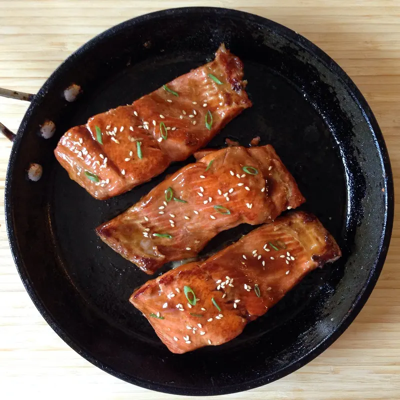 Three sticky teriyaki-glazed salmon fillets sprinkled with sesame seeds and spring onion in a black frying pan
