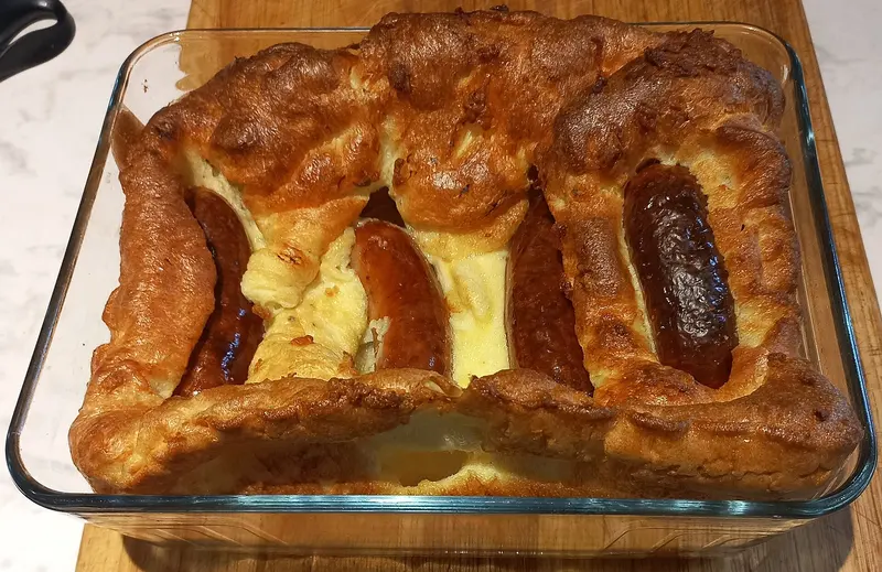 Toad in the hole in a glass baking dish, with browned sausages nestled in tall, golden puffed Yorkshire pudding batter