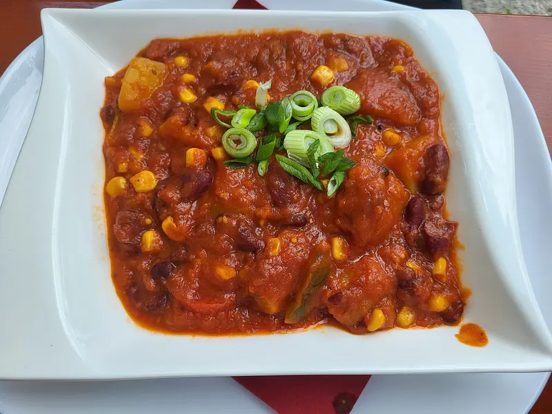 Vegan bean chilli with kidney beans, sweetcorn and chunks of potato in a rich tomato sauce, topped with spring onion, in a square white bowl