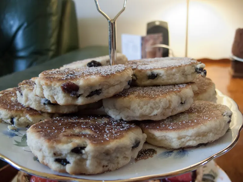 Stack of sugar-dusted Welsh cakes studded with currants, piled on a china cake stand